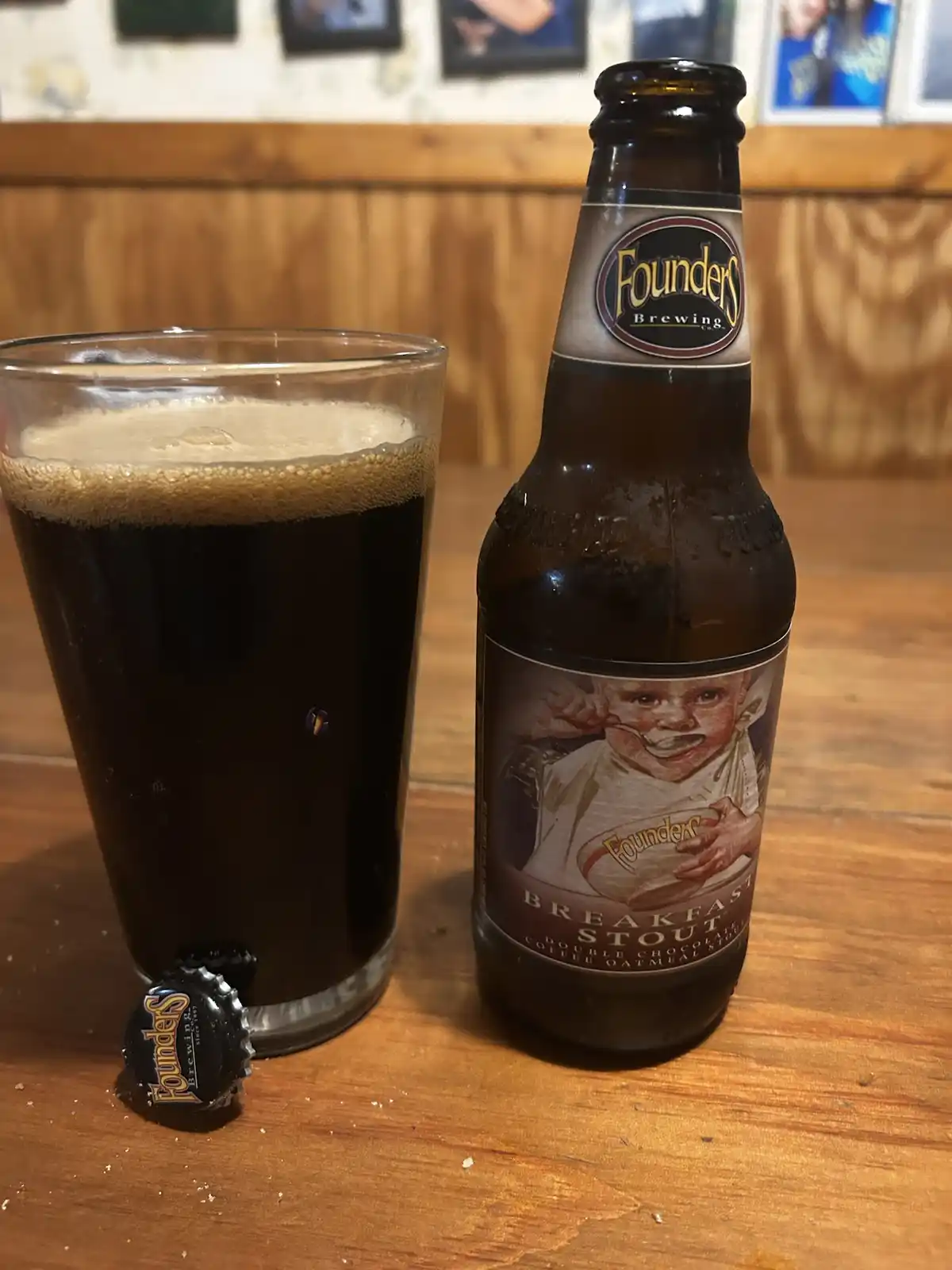 A bottle of Founders Breakfast Stout beer and a full glass sit on a wooden table, with the bottle cap beside the glass. The…