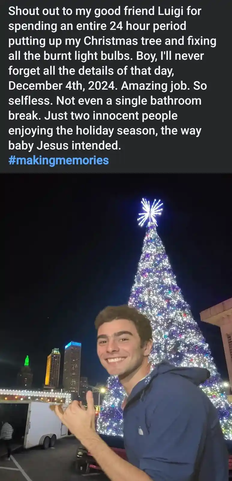 A man in a blue hoodie smiles and gives a thumbs-up in front of a large, brightly lit Christmas tree at night, with city…