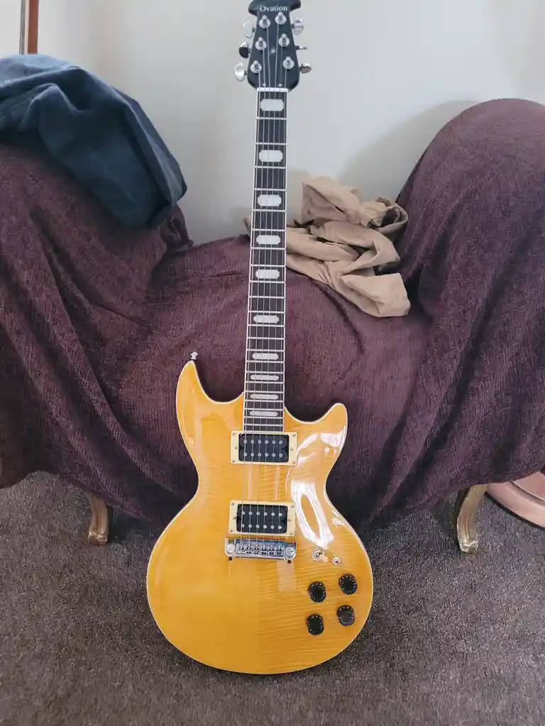 A yellow Ovation electric guitar with a glossy finish and black hardware is leaning upright against a purple upholstered…