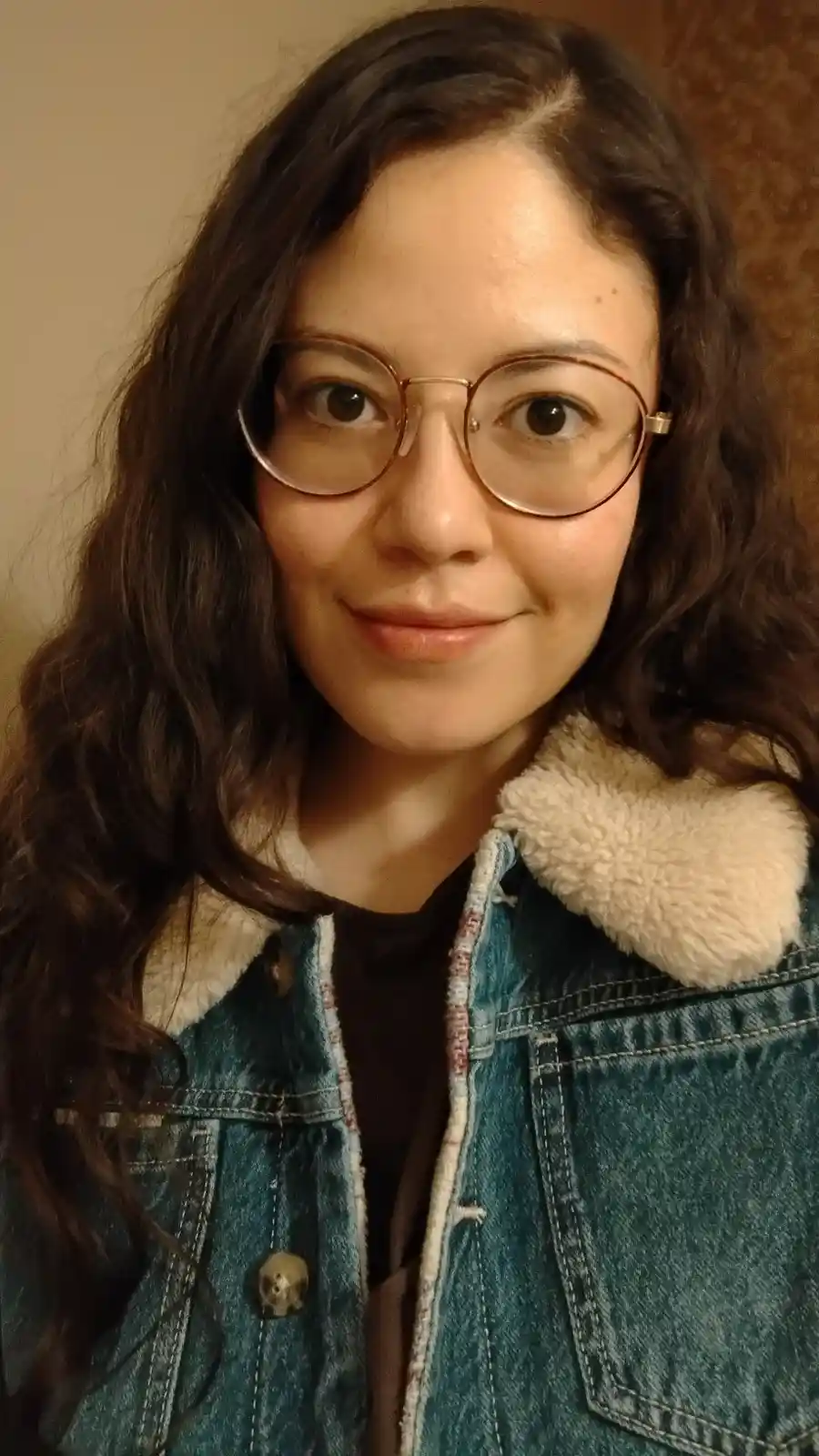 A woman with dark curly hair and round glasses smiles at the camera while wearing a denim jacket with a fluffy collar. The…