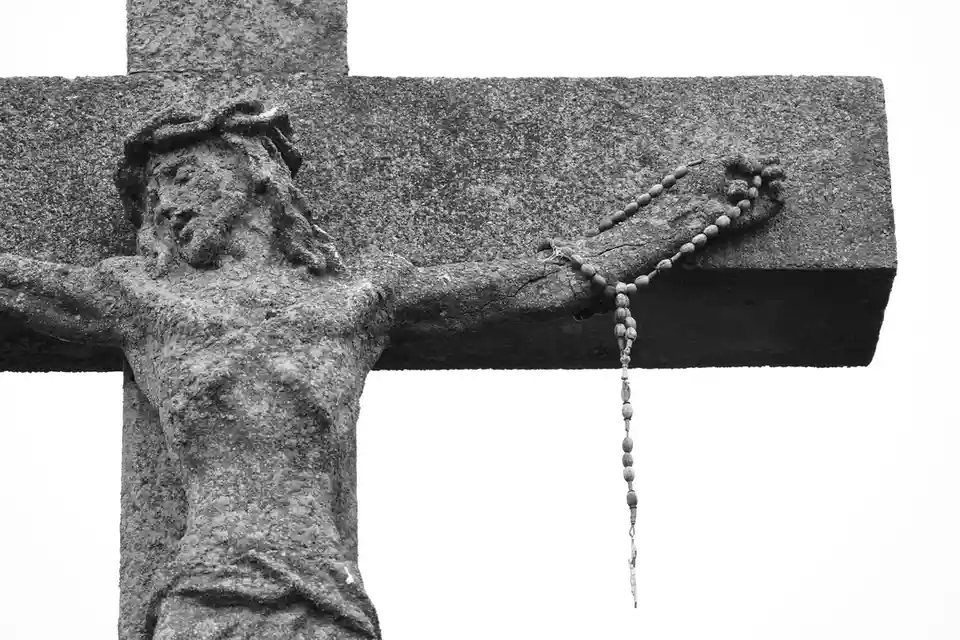 A black-and-white photograph shows a weathered stone sculpture of Jesus Christ on a cross, with a wooden rosary draped over…
