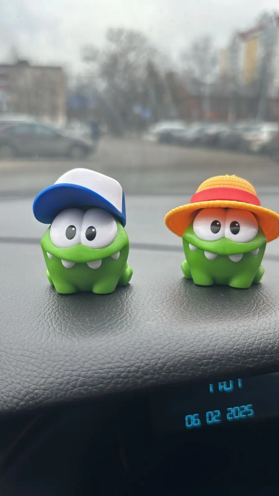 Two green, cartoonish frog-like figurines sit on a car dashboard; one wears a blue-and-white baseball cap, the other an…
