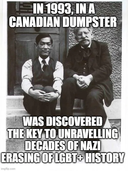 A black-and-white photo shows two men sitting side-by-side on stone steps. The man on the left is younger, wearing a vest…