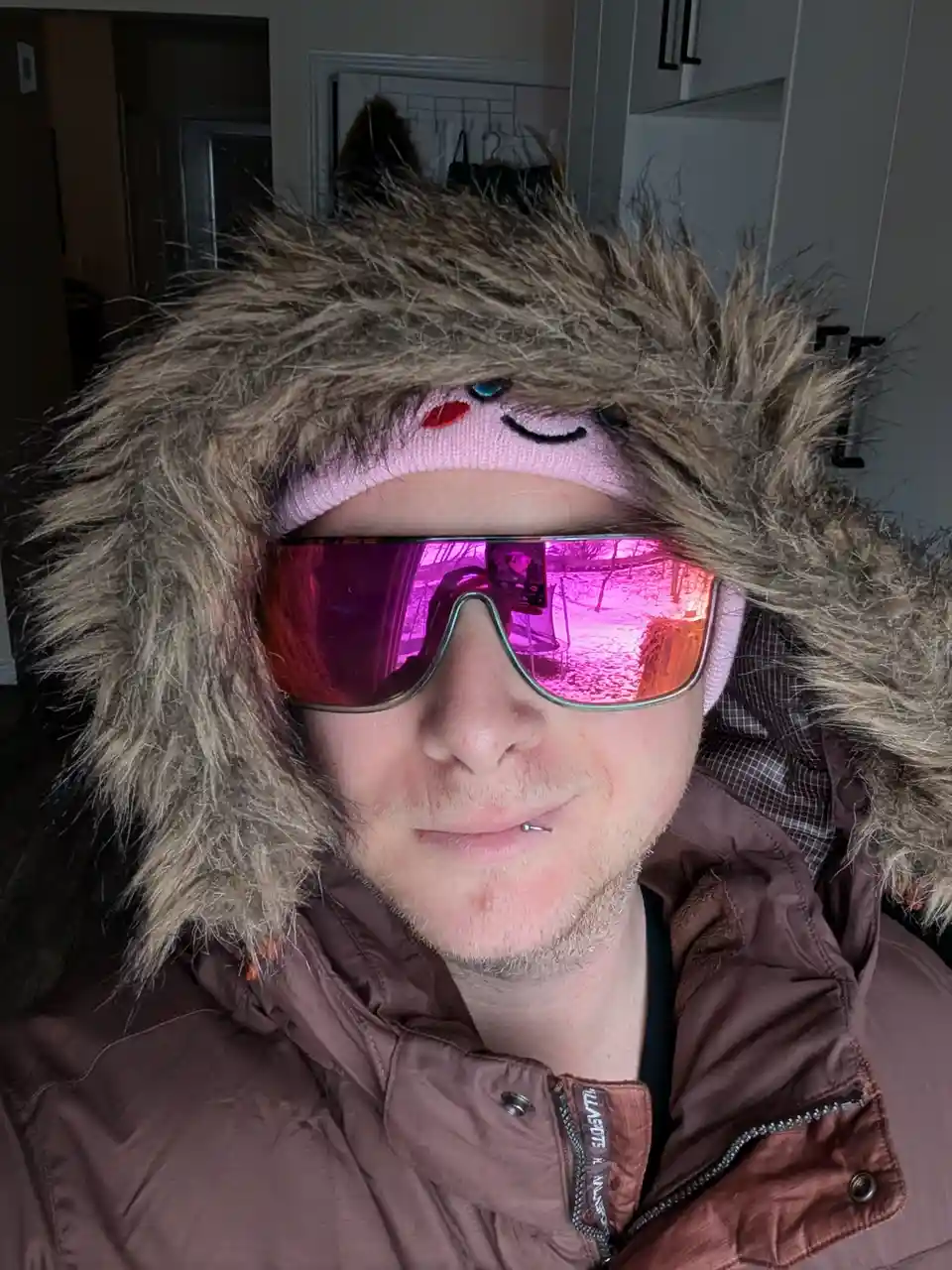 A person wearing a brown puffer jacket with a large, fluffy fur-lined hood, a pink knit hat with a cartoon face, and…