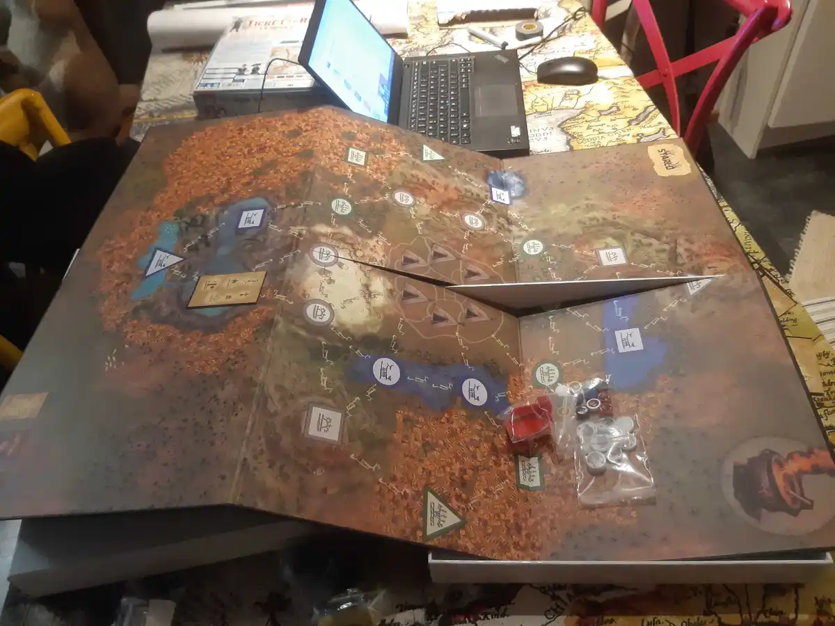 A tabletop view of a large, unfolded board game with various tokens, cards, and a laptop nearby. The board features a…