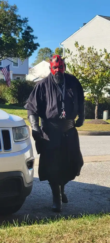 A man dressed as Darth Maul from Star Wars, complete with red mask, horns, black robe, and a lanyard with an ID badge, walks…