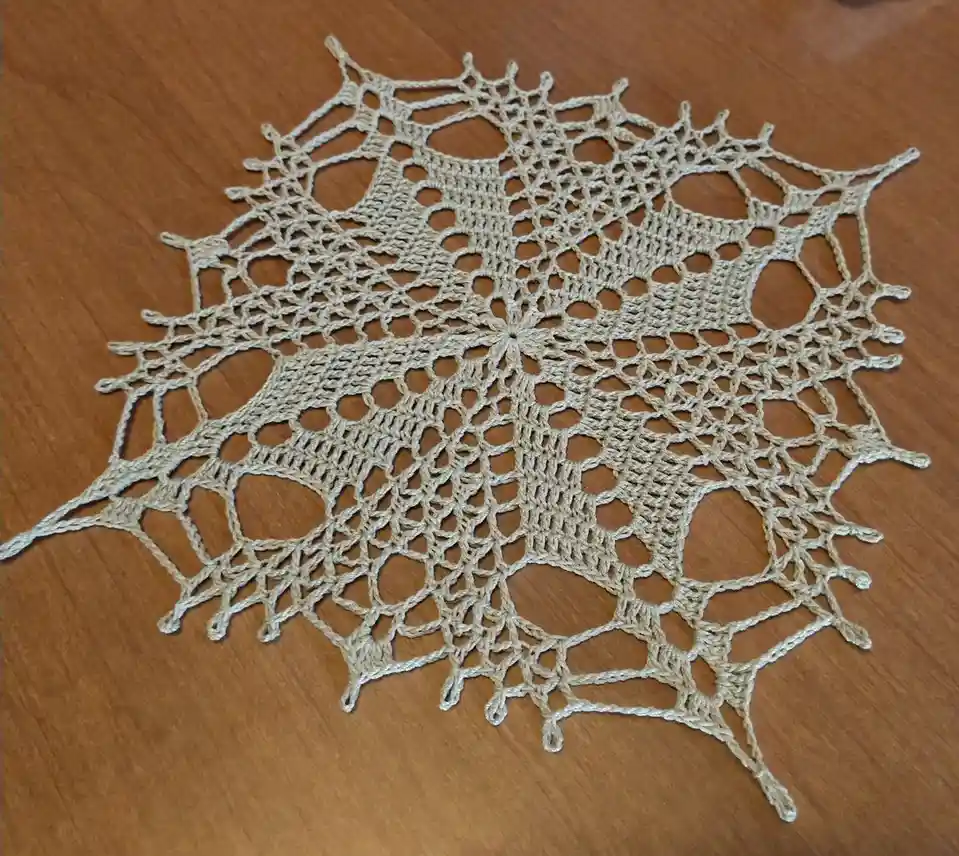 A beige, hand-crocheted doily with a spiderweb-like pattern lies on a wooden surface. The text overlaid on the image reads…