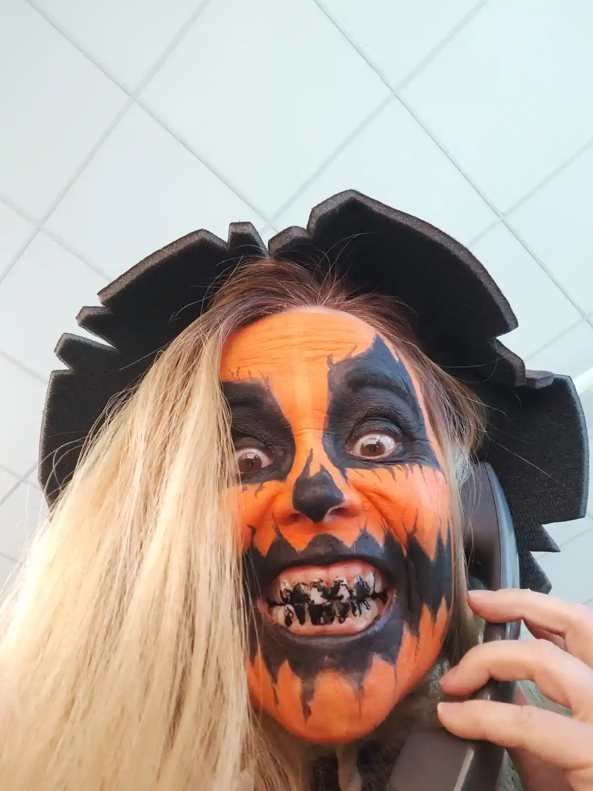 A person with long blonde hair wears elaborate orange and black Halloween pumpkin face paint and a black, jagged-edged…