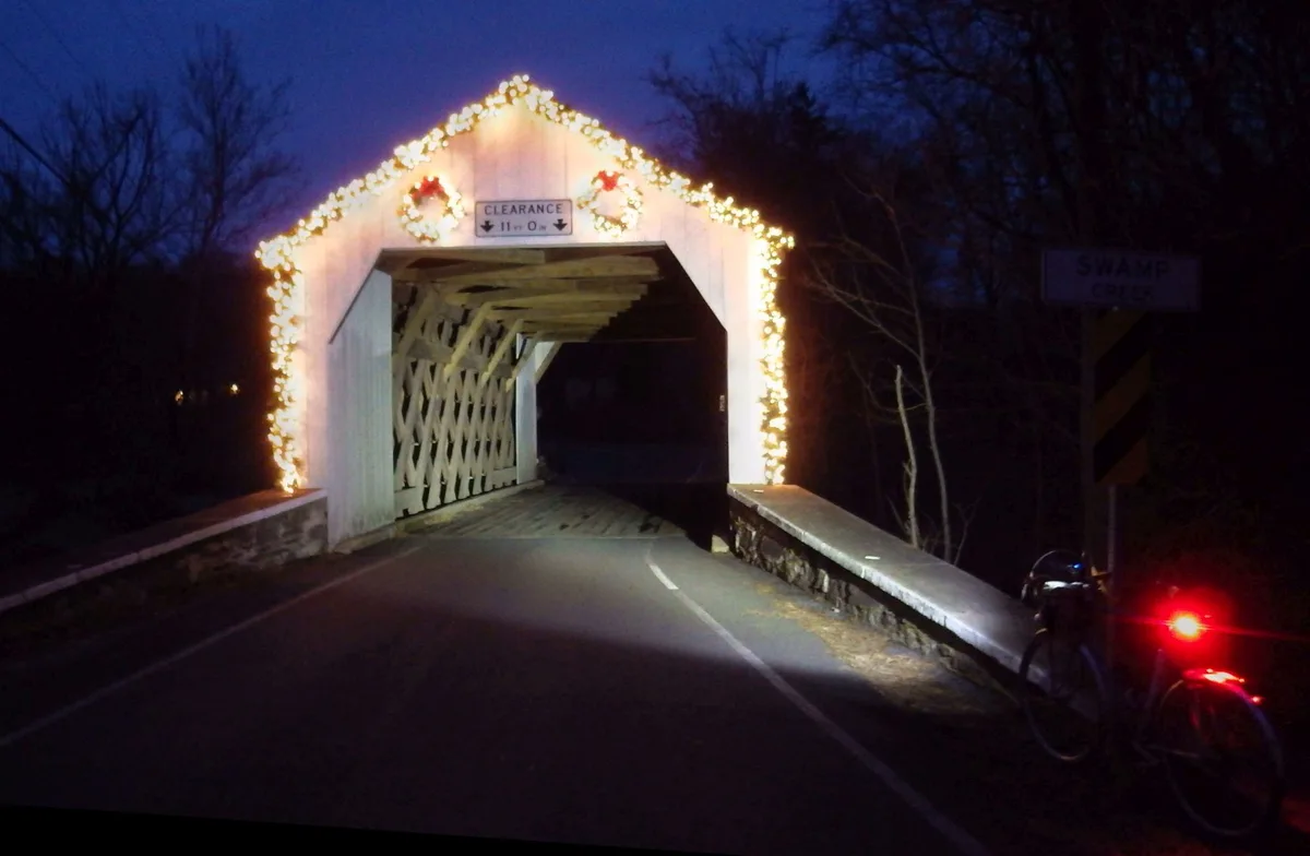 A nighttime photo shows a decorated covered bridge with festive lights and wreaths, featuring a clearance sign reading…