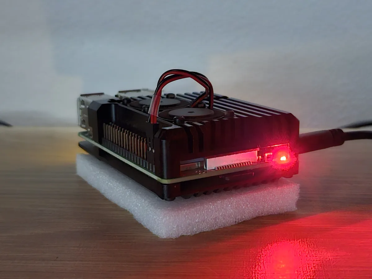 A small black computer board, resembling a Raspberry Pi, sits on a white foam pad on a wooden surface. It has a red power…