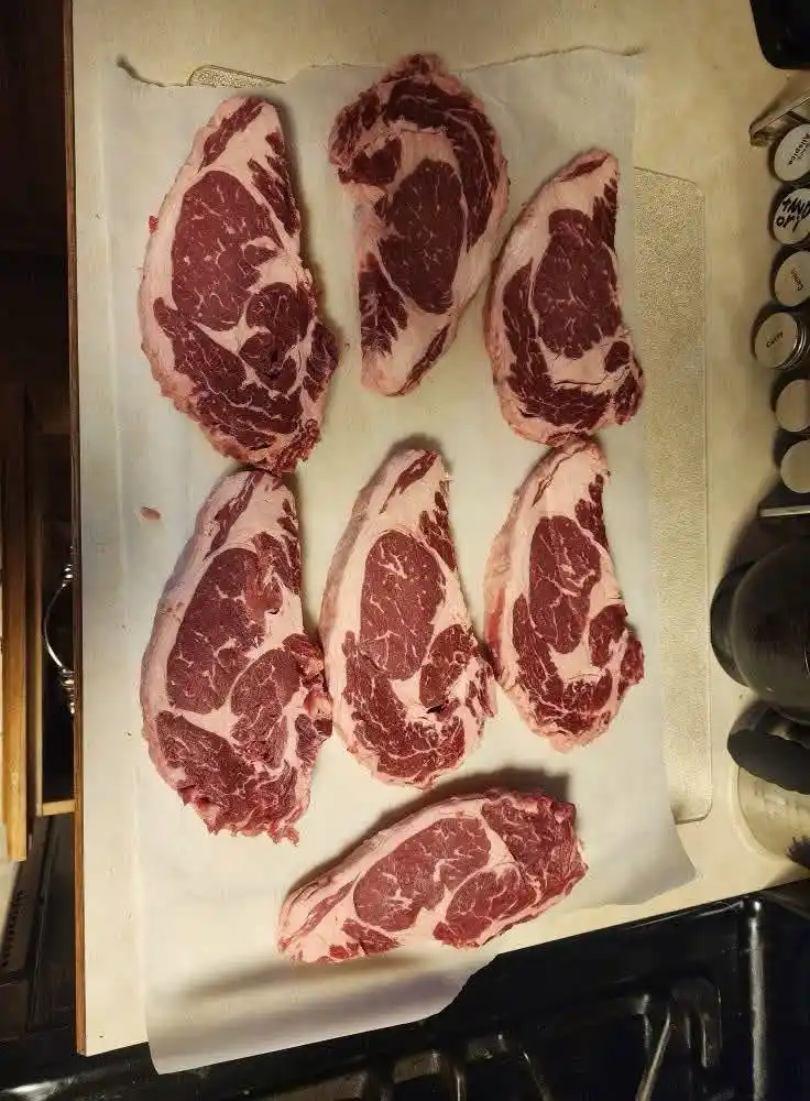 Seven raw, marbled beef steaks are arranged on parchment paper on a kitchen counter, with spice jars visible nearby. The…