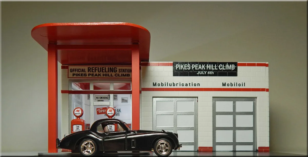 A miniature model of a vintage Mobilgas gas station with a black classic car parked at the pumps. Signs read 'OFFICIAL…