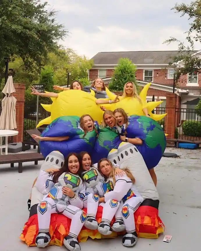 A group of young women pose together in large, inflatable costumes: three as suns with yellow rays, three as Earths with…
