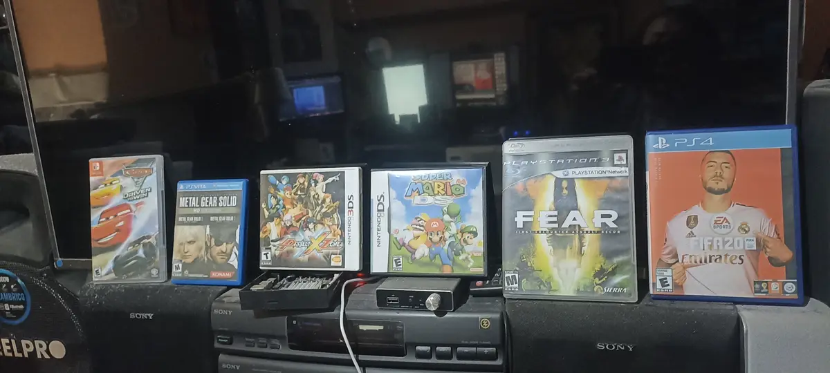 Six video game cases are lined up on top of electronic equipment, including Cars 2, Metal Gear Solid HD Collection, Project…