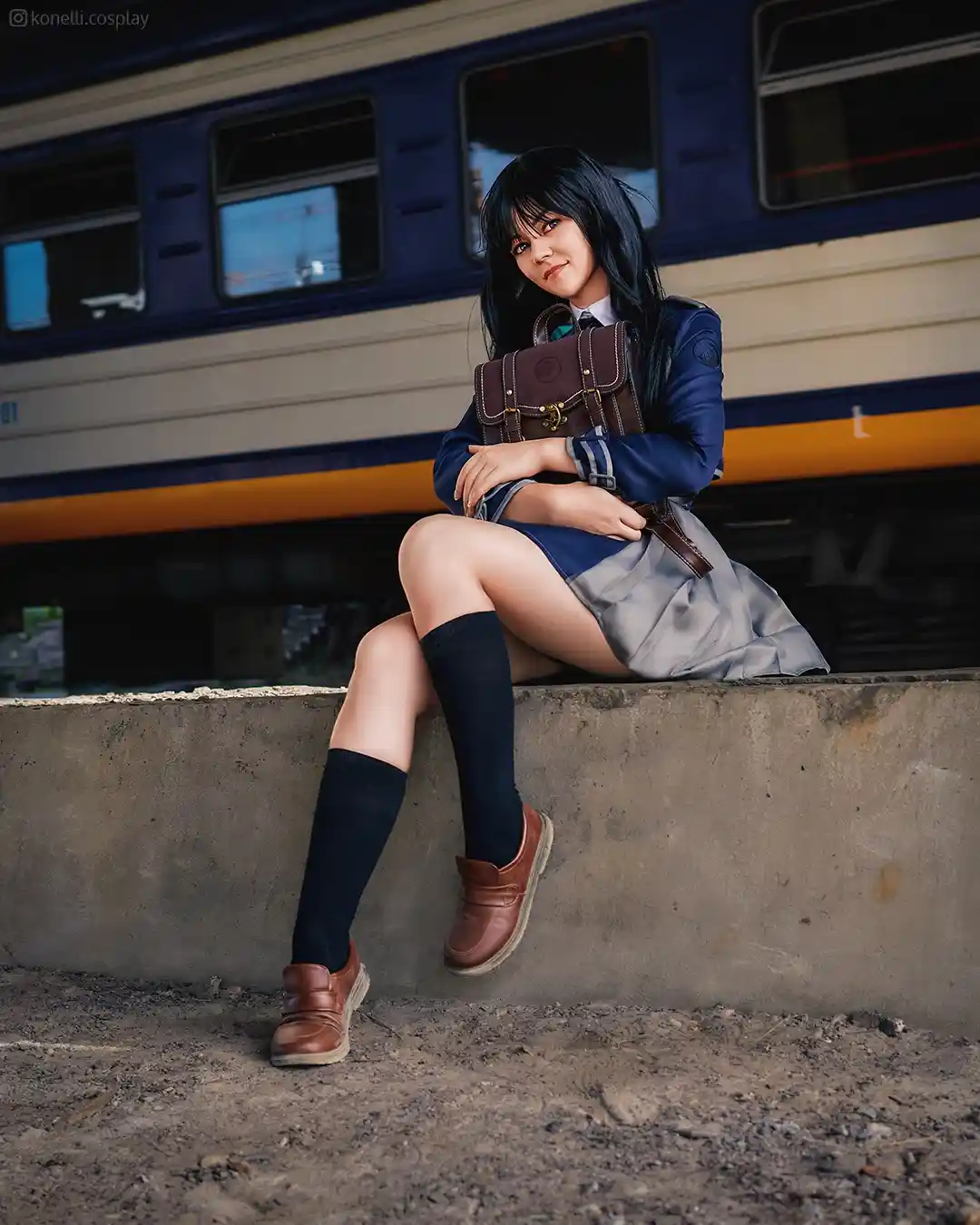 A person in a detailed school uniform cosplay, complete with a blue blazer, grey skirt, black knee-high socks, and brown…