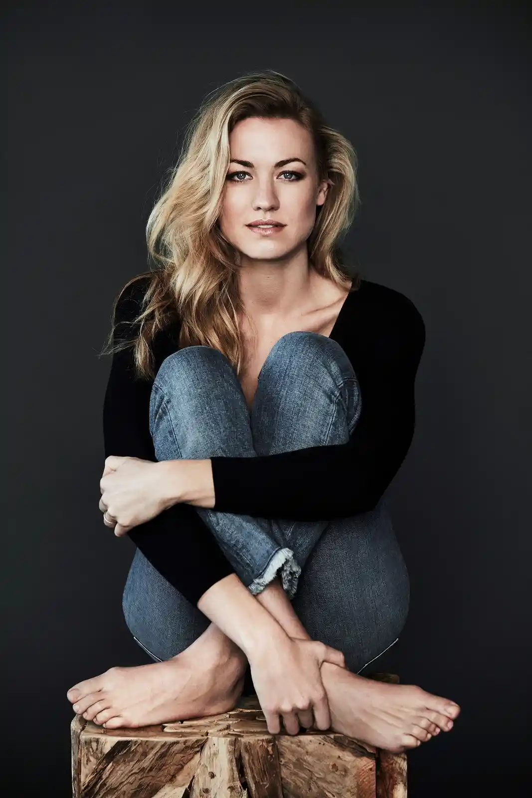 A woman with blonde hair sits barefoot on a wooden stump, wearing a black long-sleeved shirt and blue jeans, hugging her…