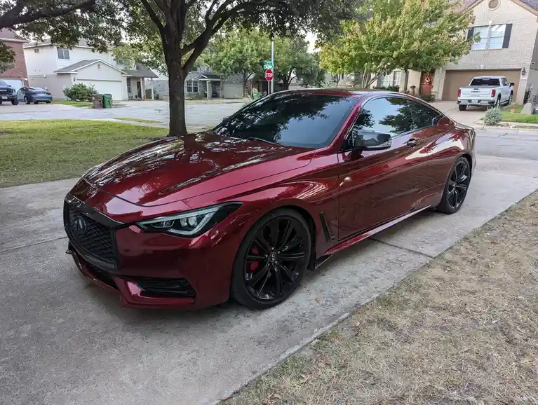 A glossy red Infiniti Q60 sports coupe is parked on a residential driveway, featuring black wheels and a sleek design. The…