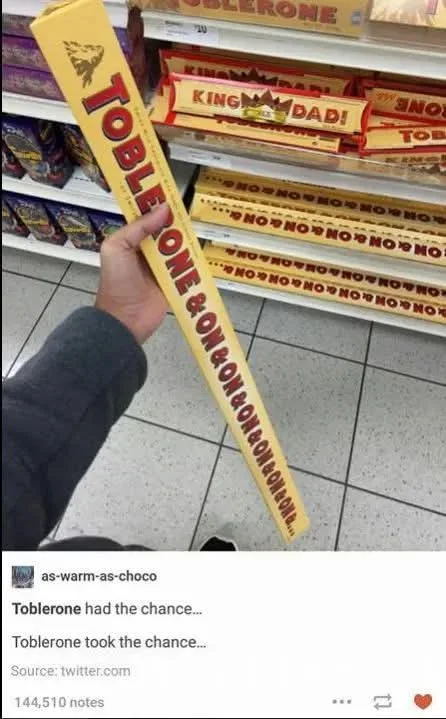 A person holds a very long, unusually oversized Toblerone chocolate bar in a store aisle, with shelves of regular Toblerone…
