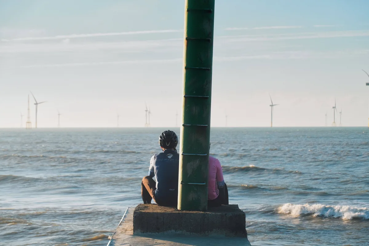 Two cyclists in cycling gear sit back-to-back on a concrete pier, partially obscured by a green pole, looking out at a body…