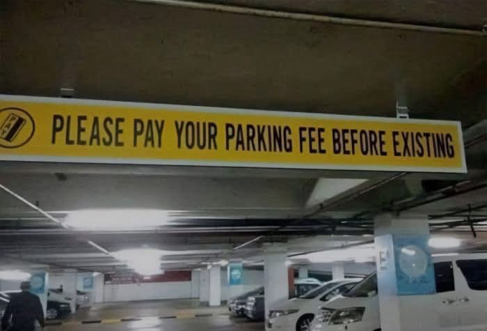 A yellow overhead sign in a dimly lit parking garage reads 'PLEASE PAY YOUR PARKING FEE BEFORE EXISTING'. The sign features…
