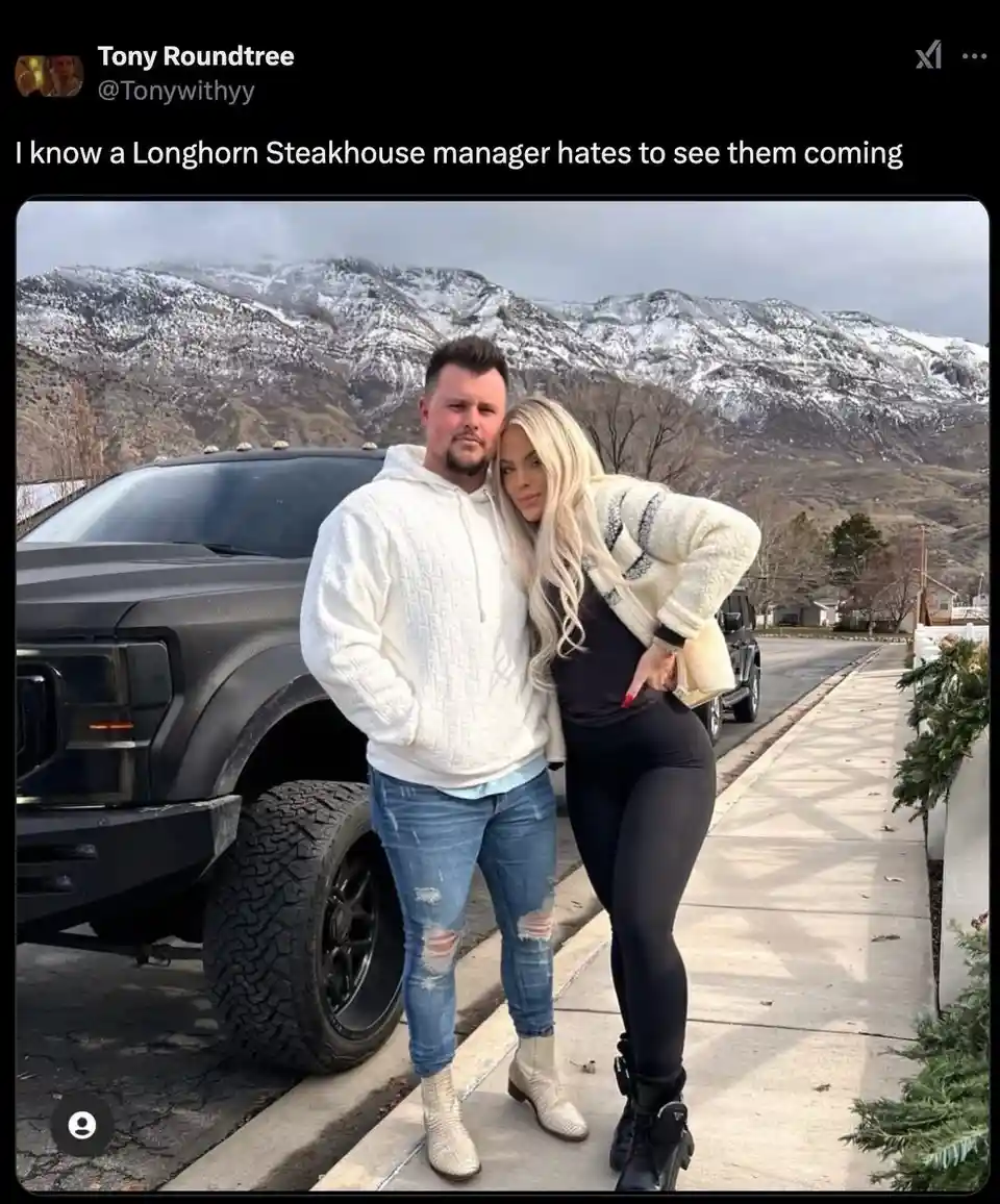 A man and a woman pose together on a sidewalk next to a large black truck, with snow-capped mountains in the background. The…