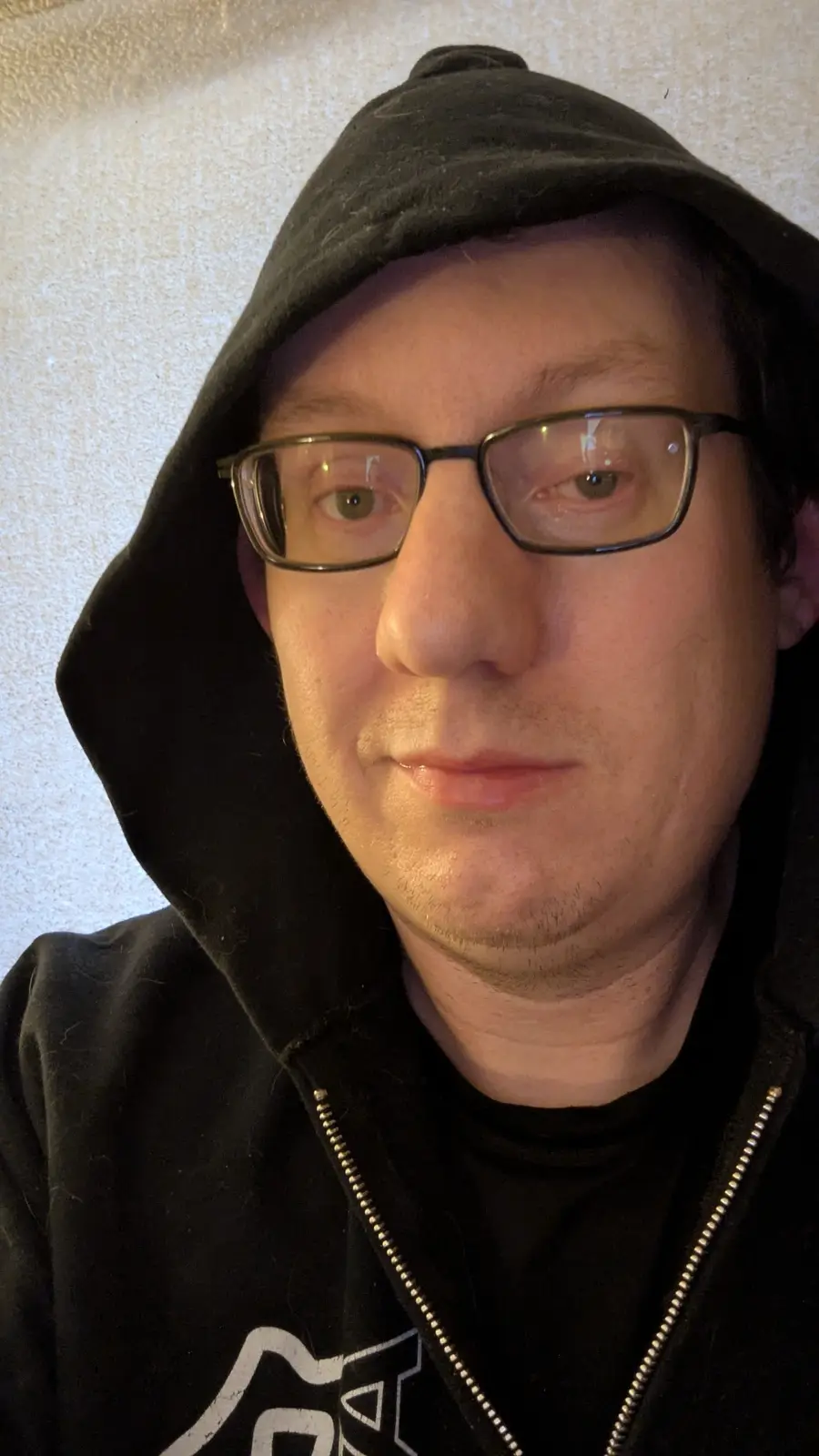 A close-up selfie of a man wearing a black hoodie with the hood up and black-rimmed glasses, looking directly at the camera…
