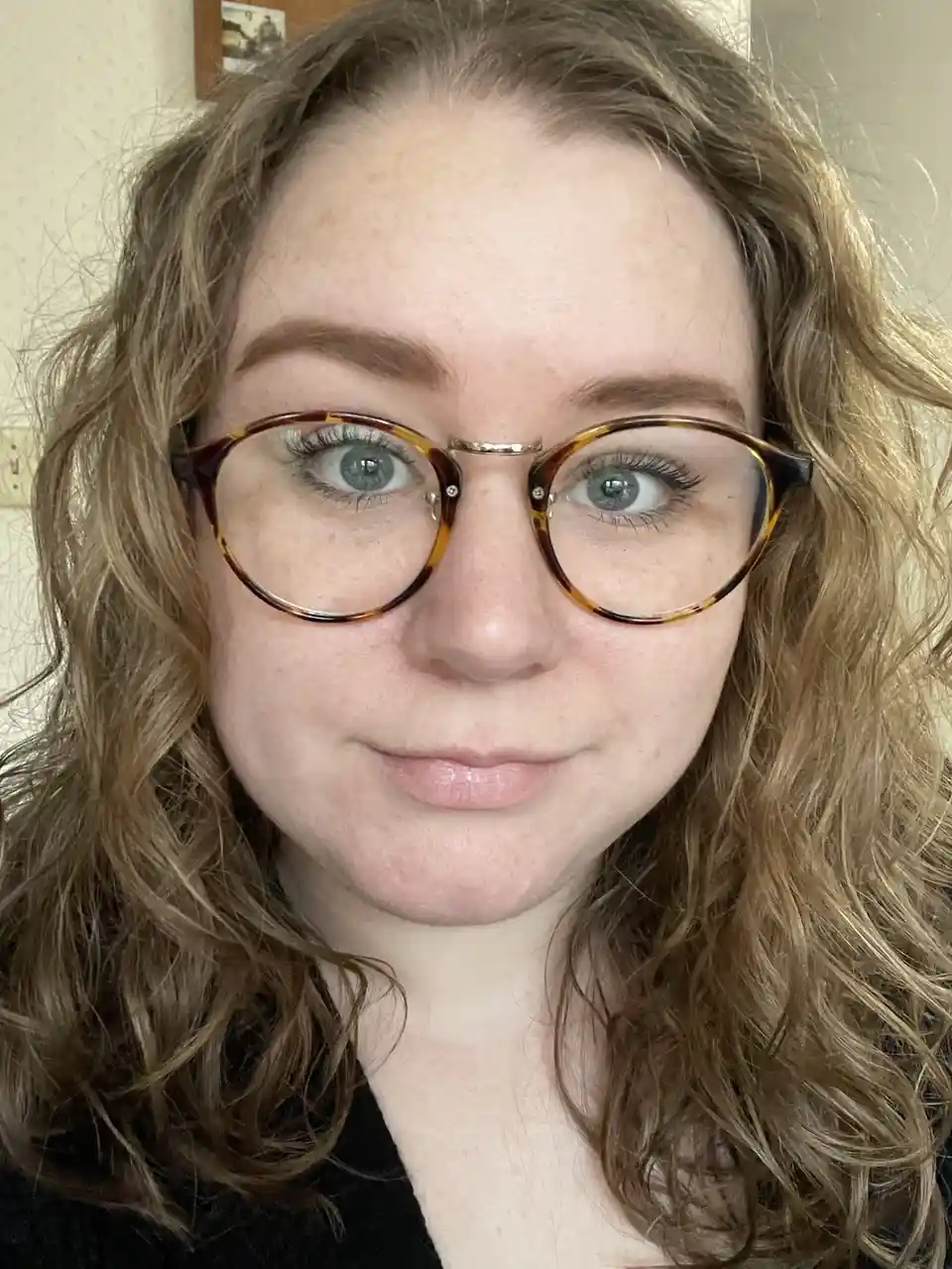 A close-up selfie of a person with curly brown hair and tortoiseshell glasses, looking directly at the camera with a slight…