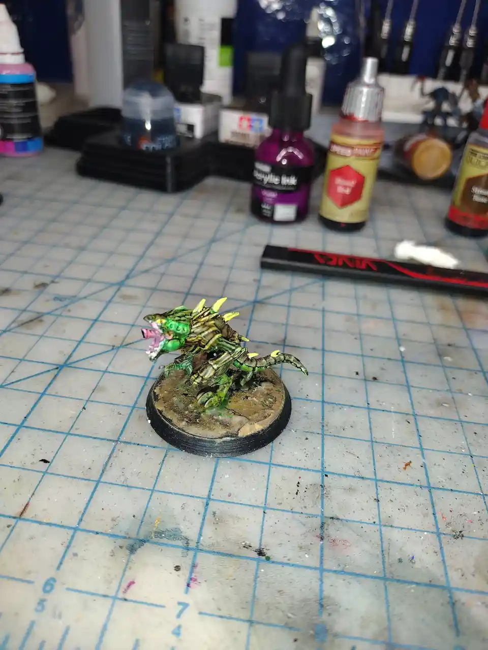 A painted miniature monster figurine with green and yellow spikes, standing on a circular base, is centered on a blue grid…
