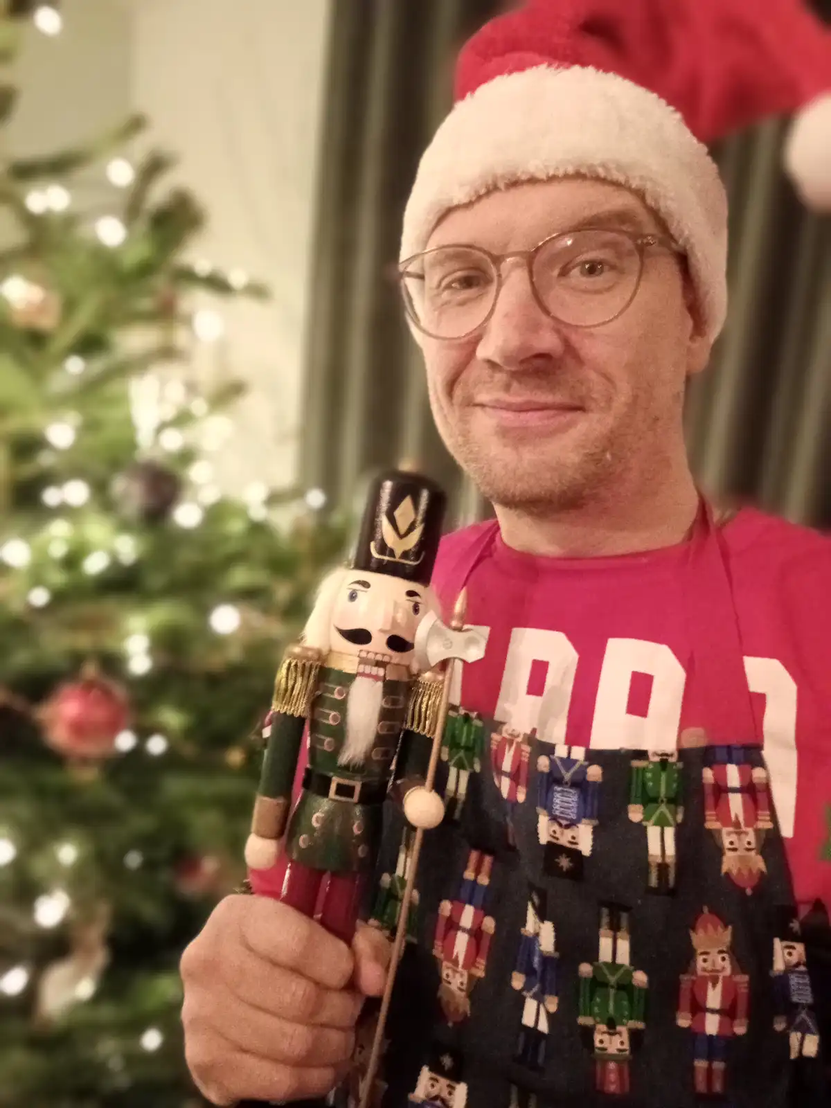 A man wearing a red Santa hat and a festive sweater covered in nutcracker patterns holds a large wooden nutcracker figurine…