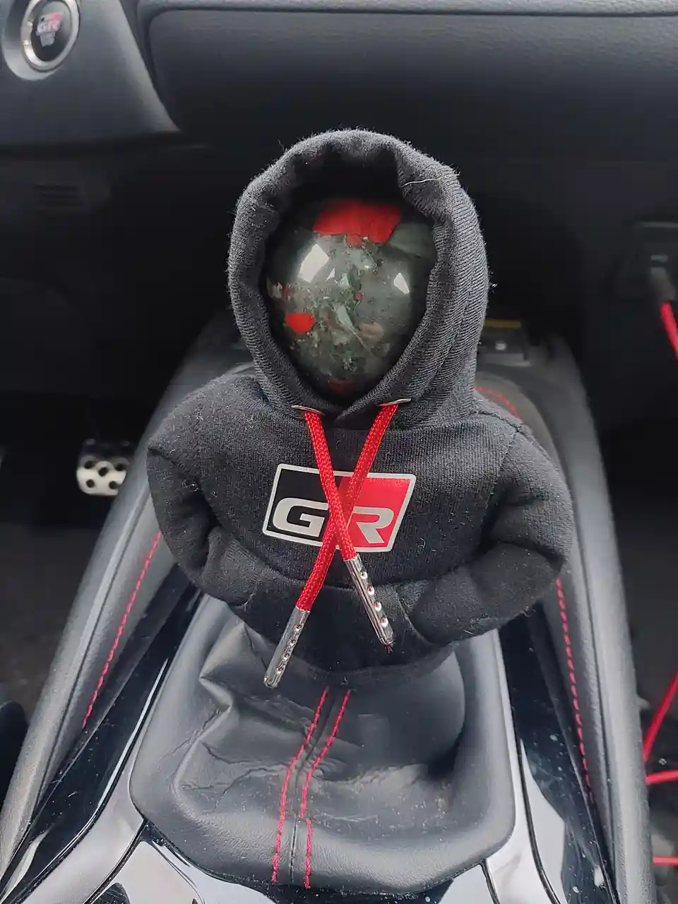A close-up shot of a car's manual gear shifter knob, which is covered by a small black hoodie with red drawstrings and a GR…