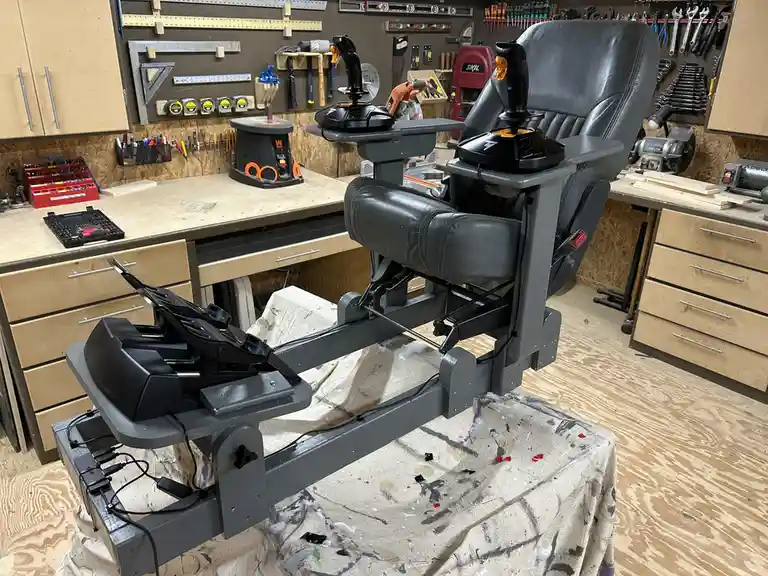 A custom-built flight simulator chair with two joysticks and foot pedals is mounted on a grey metal frame in a cluttered…