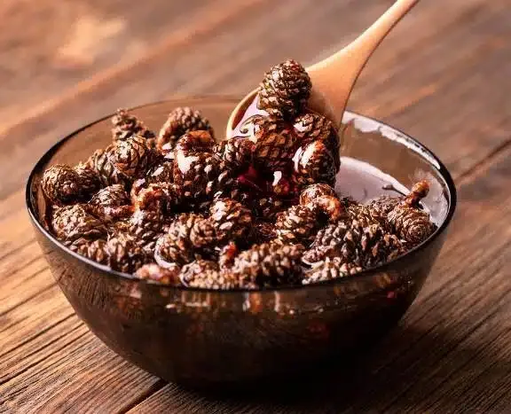 A dark glass bowl filled with pine cones submerged in a dark, glossy liquid, with a wooden spoon lifting several pine cones…