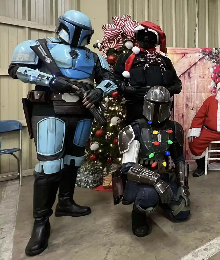 Three people in Mandalorian armor cosplay pose in front of a decorated Christmas tree. The standing figure wears blue armor…