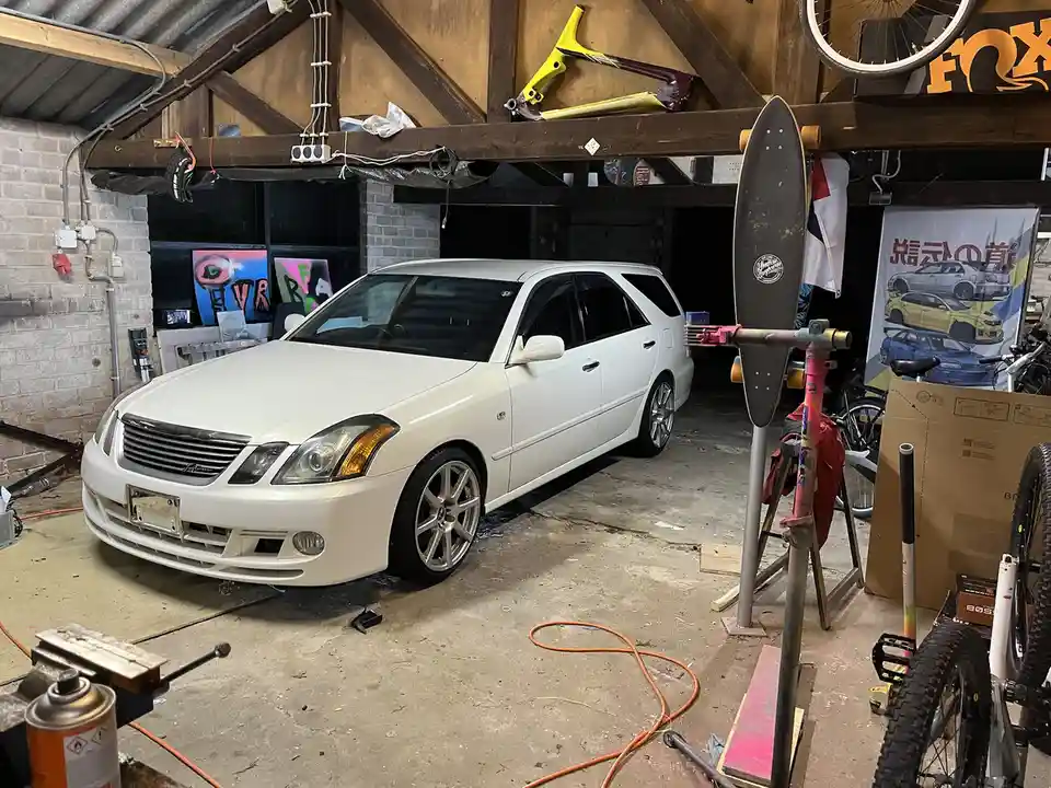 a white toyota crown station wagon is parked inside a cluttered garage filled with various bikes, skateboards, and tools…