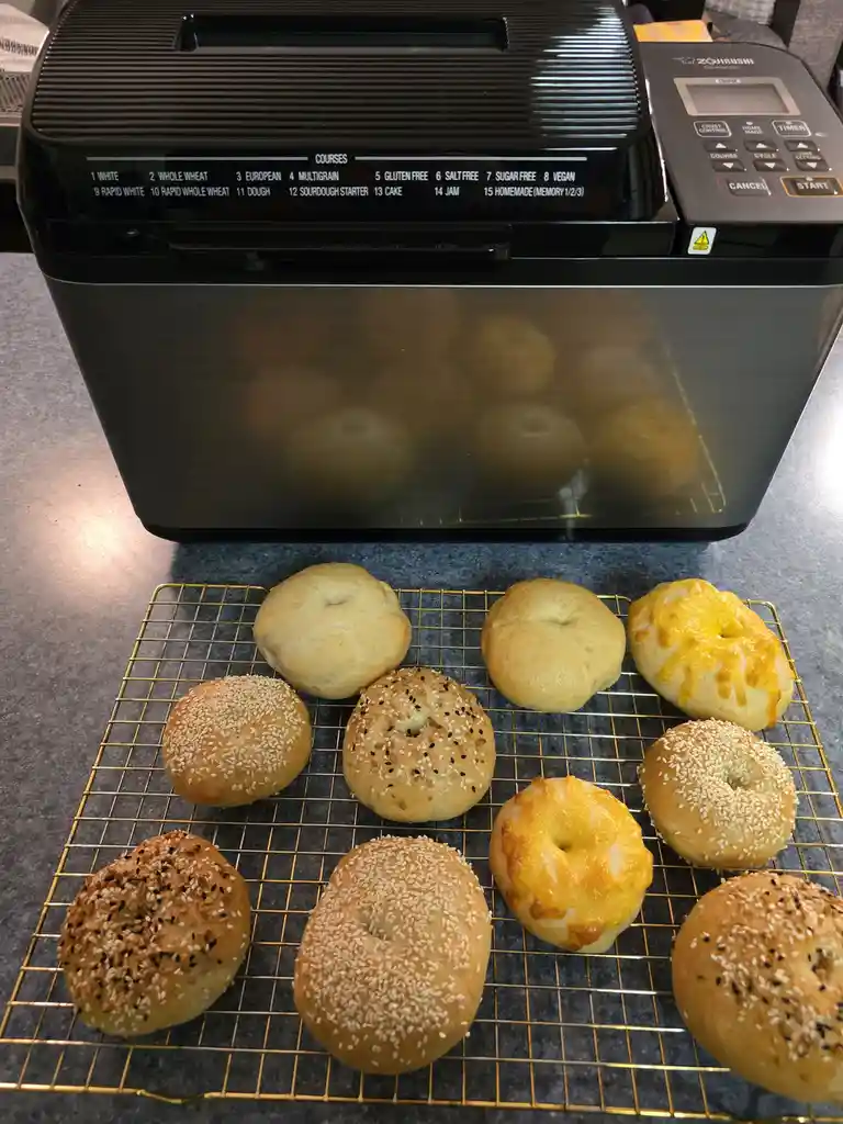 A black Zojirushi bread machine with a list of 15 preset courses is in the background, while in front of it, on a gold wire…