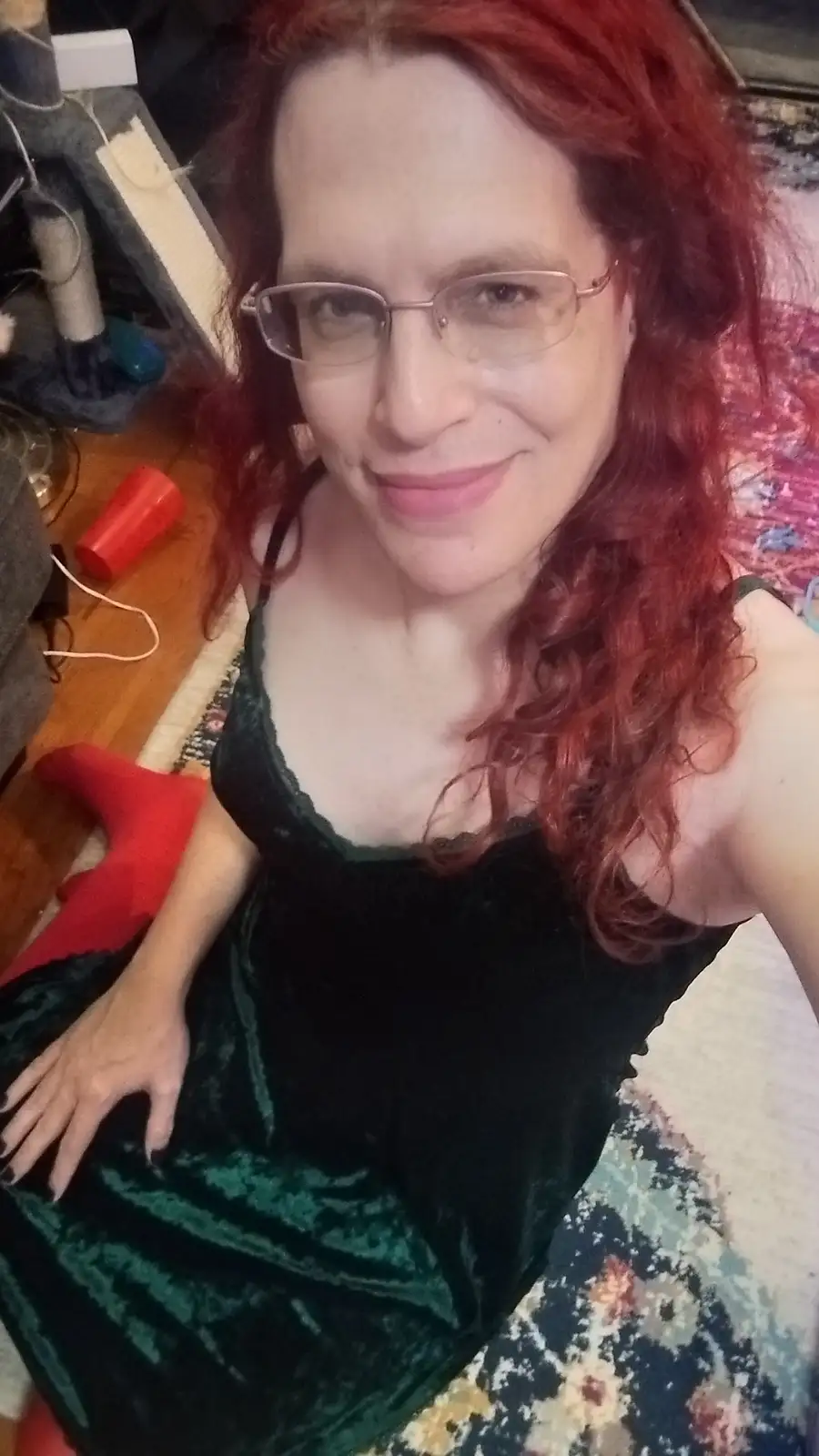 A woman with red curly hair and glasses, wearing a dark green velvet dress, smiles at the camera while seated on a patterned…