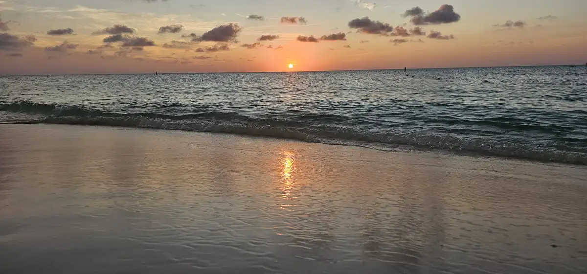A serene beach sunset with the sun low on the horizon, casting orange and pink hues across the sky and reflecting on the wet…