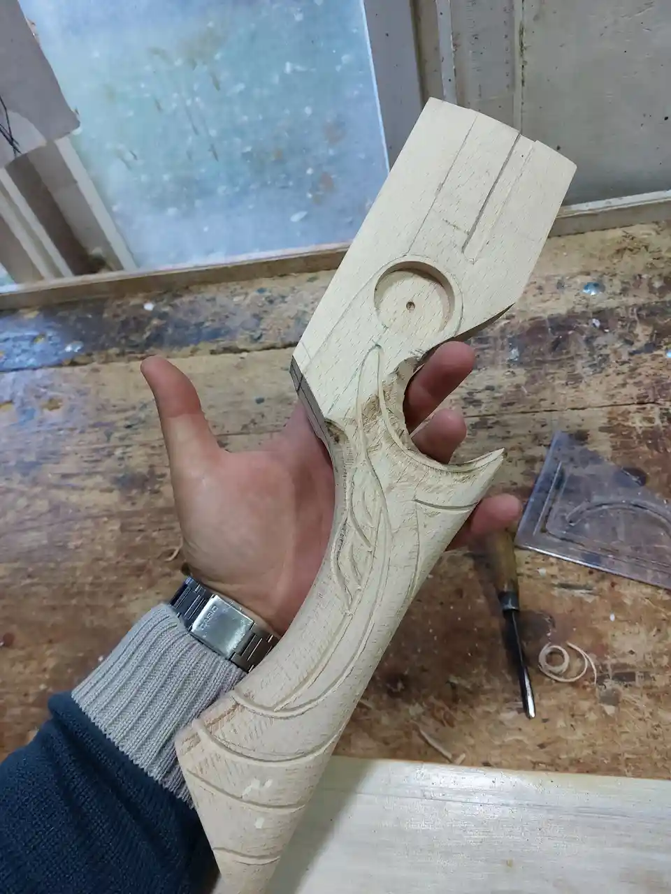 A person's hand holds a wooden carving that resembles a gun, with detailed carvings and a circular hole near the top. The…