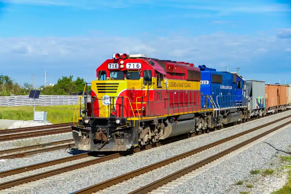 A red and yellow Florida East Coast locomotive numbered 716 leads a freight train, with the number 716 prominently displayed…
