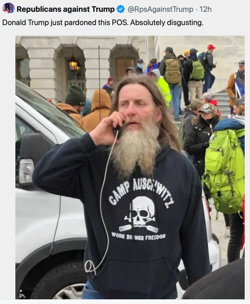 A man with a long beard and dark hoodie featuring a skull graphic and the text 'CAMP AUSCHWITZ' and 'WORK BRINGS FREEDOM' is…
