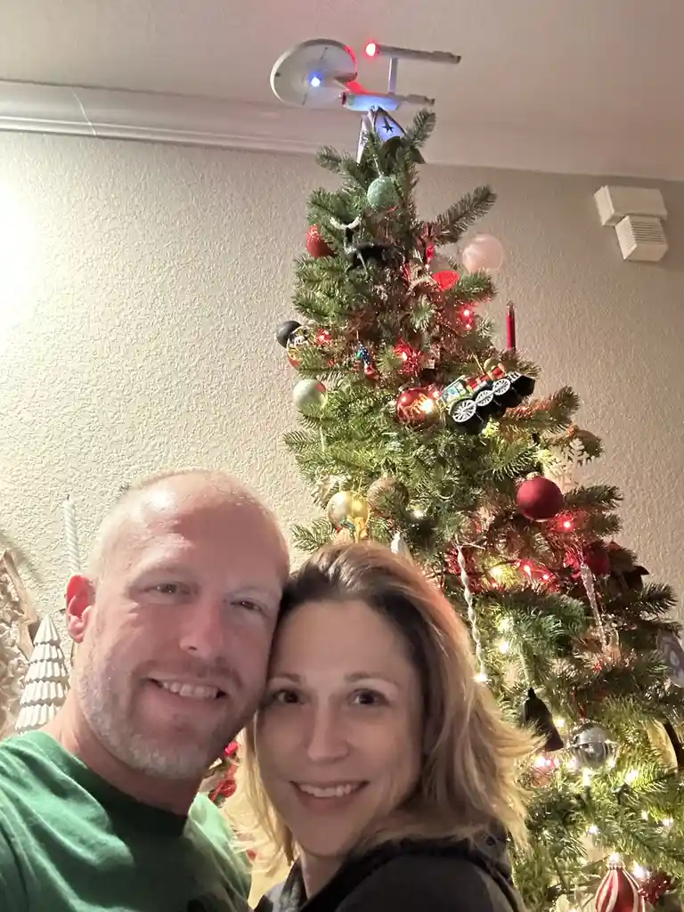 A smiling couple poses for a selfie in front of a decorated christmas tree. The tree is adorned with red and gold ornaments…