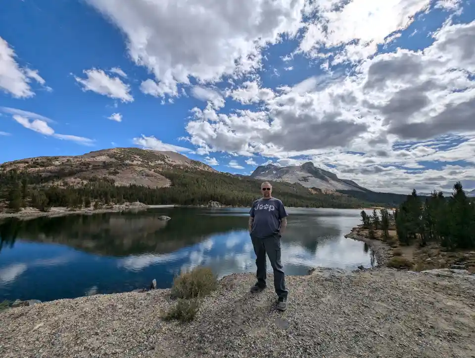 A man stands on a gravel shore beside a calm lake with mountains and a partly cloudy sky behind him. He wears a dark blue…