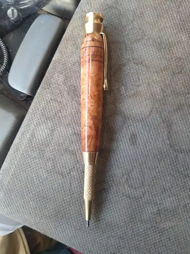 A custom-made pen with a burl wood barrel and brass accents lies on a gray textured fabric surface, with overlaid text…