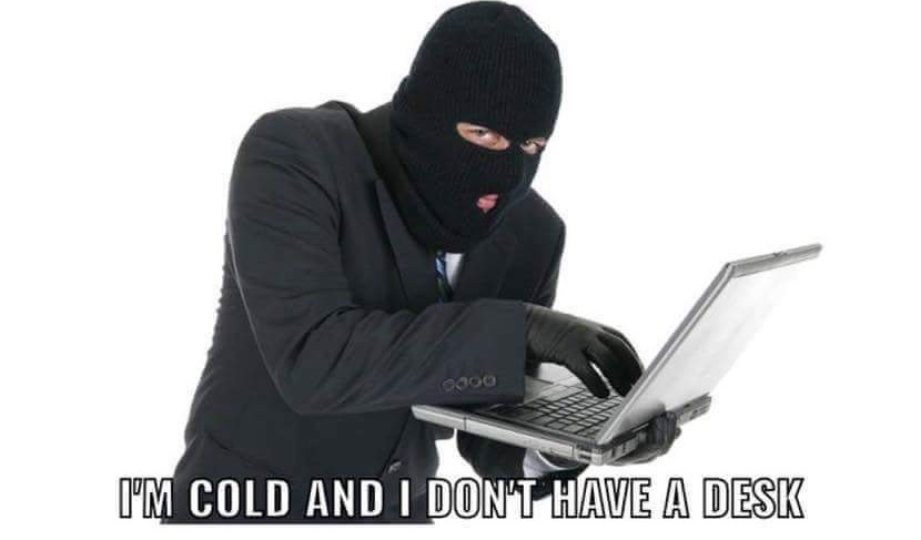 A person wearing a black ski mask and black gloves, dressed in a black suit, is hunched over a silver laptop they're holding…