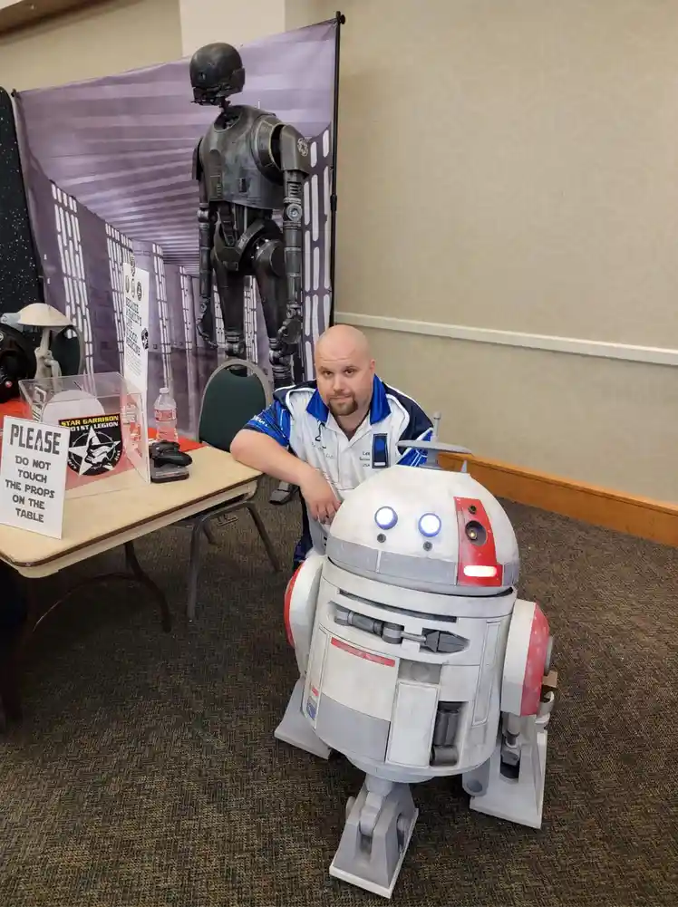 A man in a Star Wars-themed shirt poses beside a life-sized R2-D2 droid prop at a convention. Behind him, a large poster of…