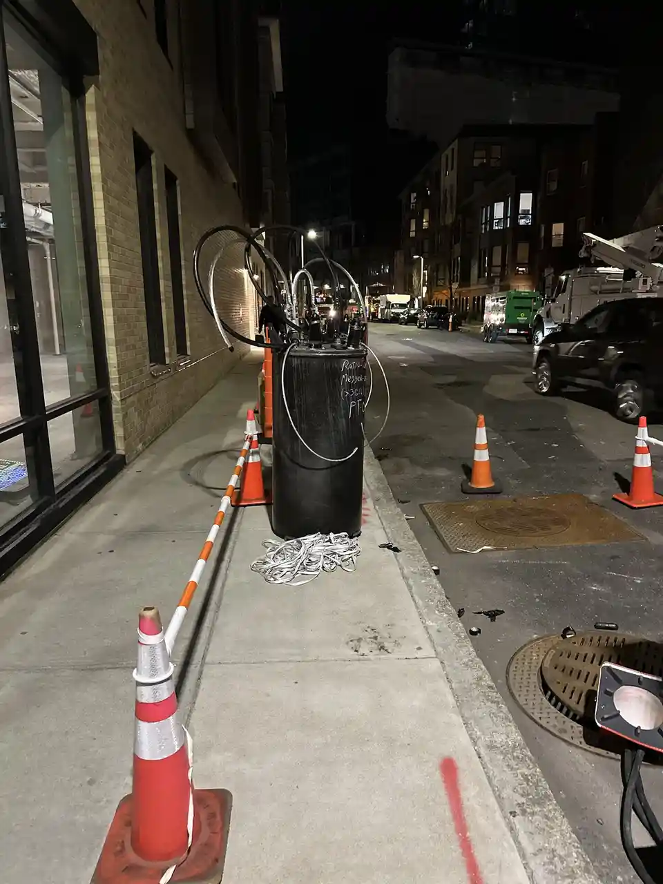 A nighttime street scene shows a black cylindrical container on a sidewalk, surrounded by orange traffic cones and tangled…