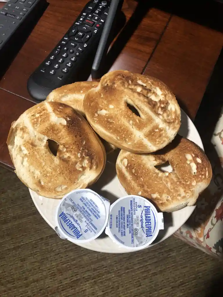 A white plate holds four toasted bagels with golden-brown, slightly burnt surfaces, accompanied by two small containers of…