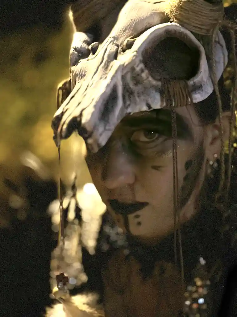 A person with dark makeup and dreadlocks wears a large, weathered animal skull as a headpiece, with strings and beads…
