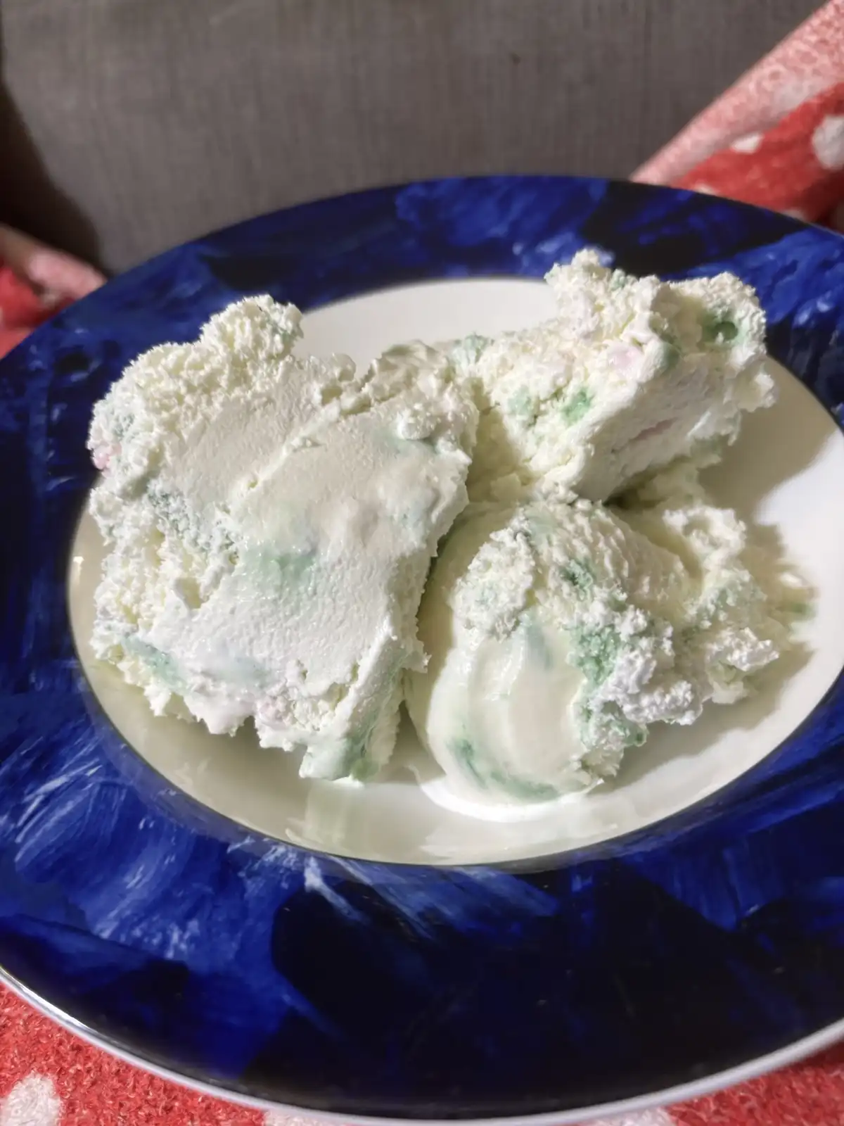 A close-up photo shows three scoops of white, textured frozen dessert with green and pink flecks on a blue-rimmed white…