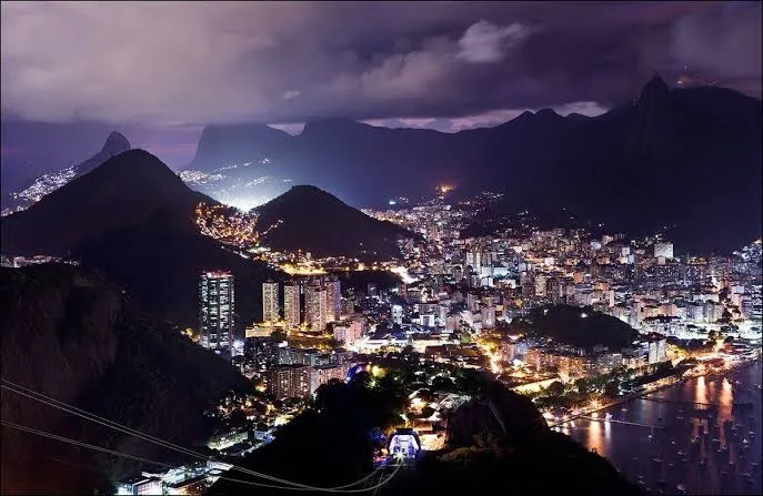 A nighttime panoramic photo of Rio de Janeiro, showing illuminated city buildings nestled between dark mountains and a…
